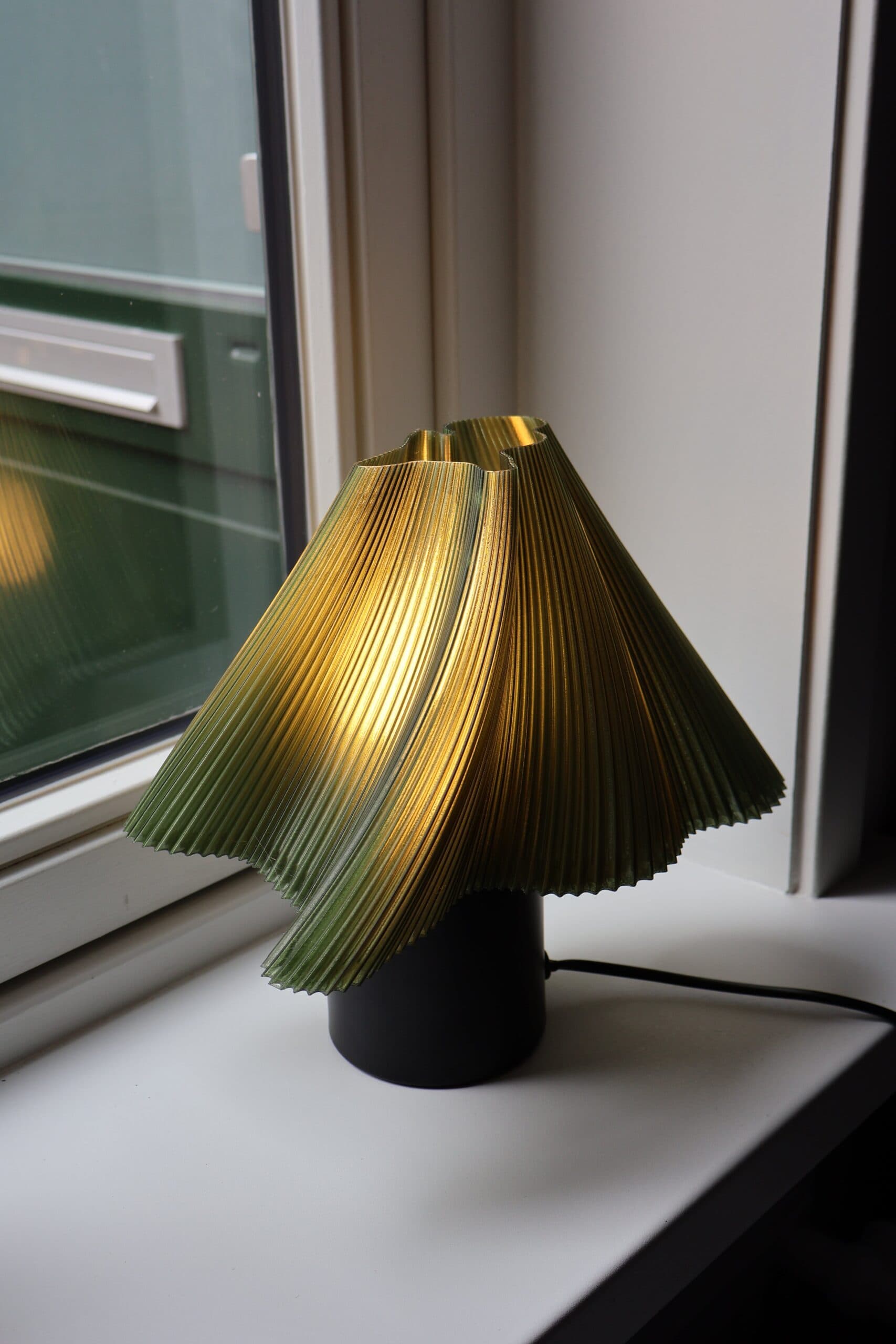 Groene tafellamp met 3D-geprinte kap op een houten vensterbank, verspreidt warm licht en creëert een gezellige sfeer.
